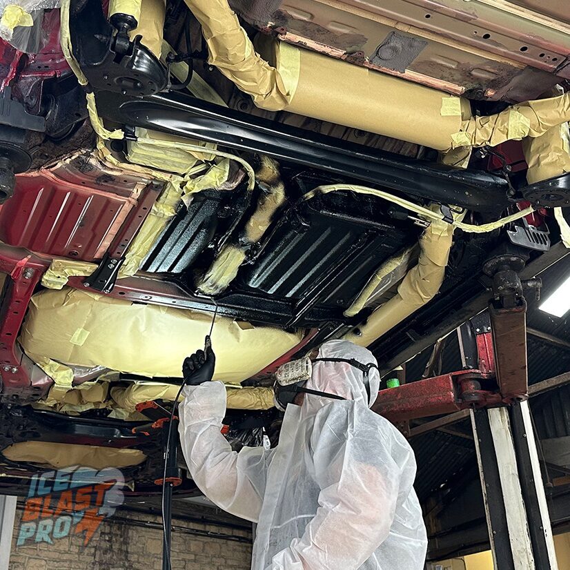 Technician in a protective suit applying Dinitrol corrosion protection to a masked vehicle underbody