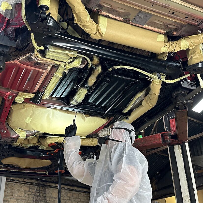 Technician in a protective suit applying Dinitrol corrosion protection to a masked vehicle underbody