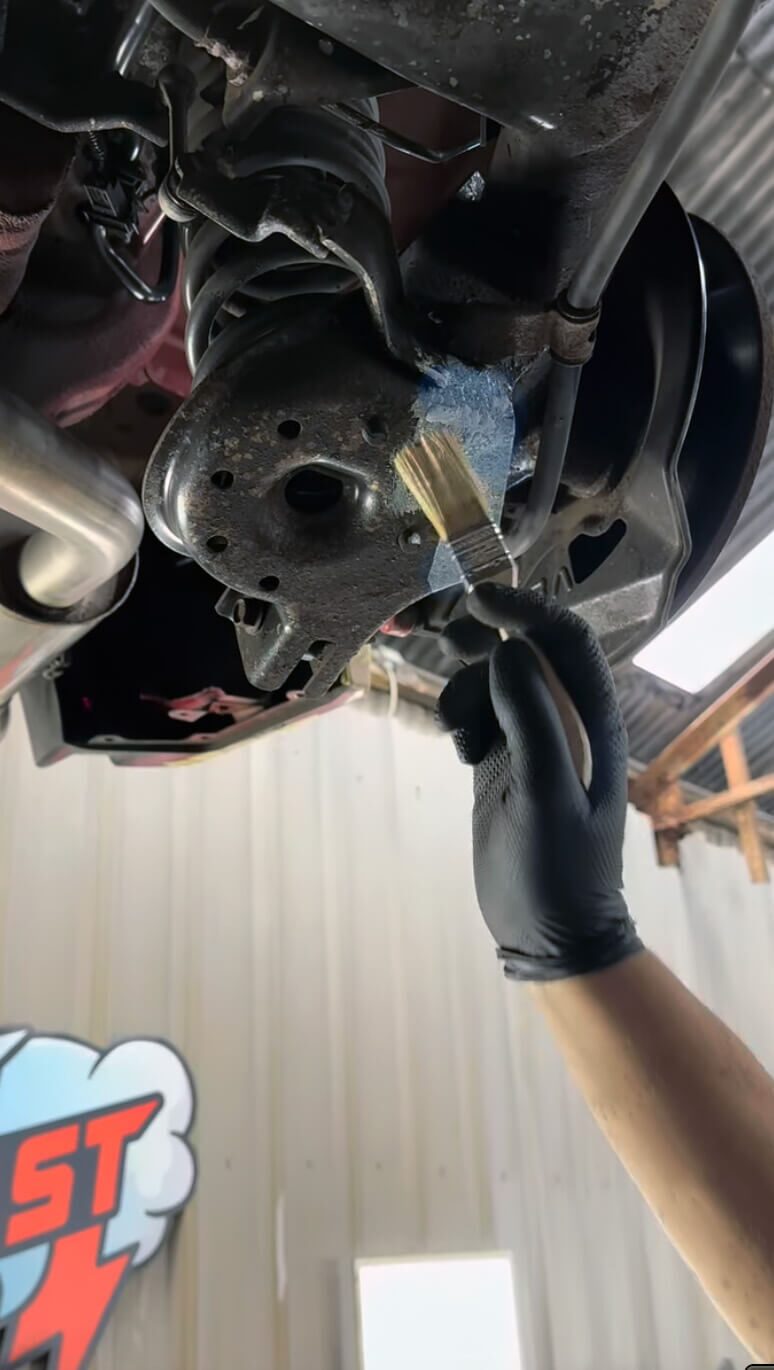 Gloved hand brushing blue chemical rust stabiliser onto a cleaned suspension component on a raised vehicle underside