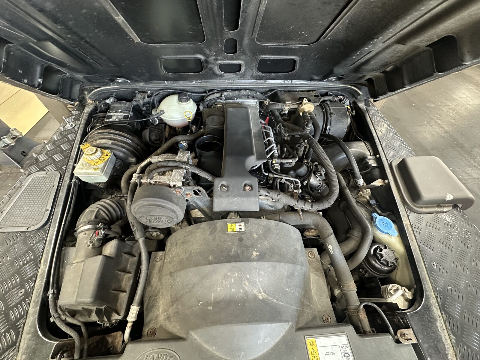 Land Rover Defender 90 engine bay before cleaning, with dust and dry grime over the engine cover, hoses and ancillaries