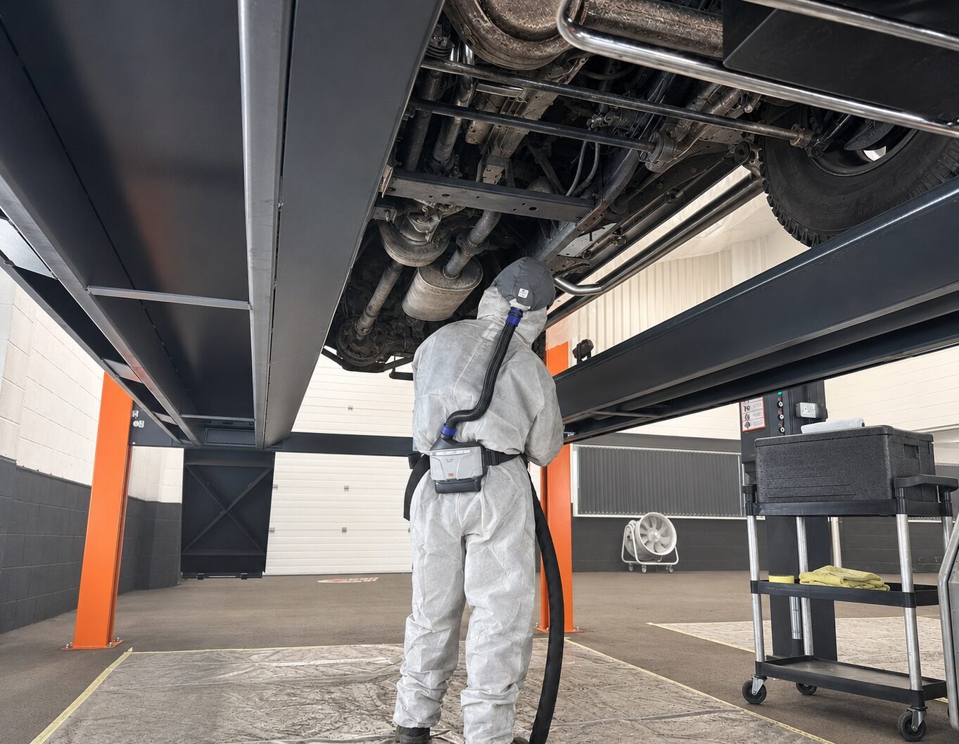 Underside of a raised 4x4 at IceBlastPro, a technician in a protective suit dry ice blasting the chassis rails, exhaust and rear axle from below