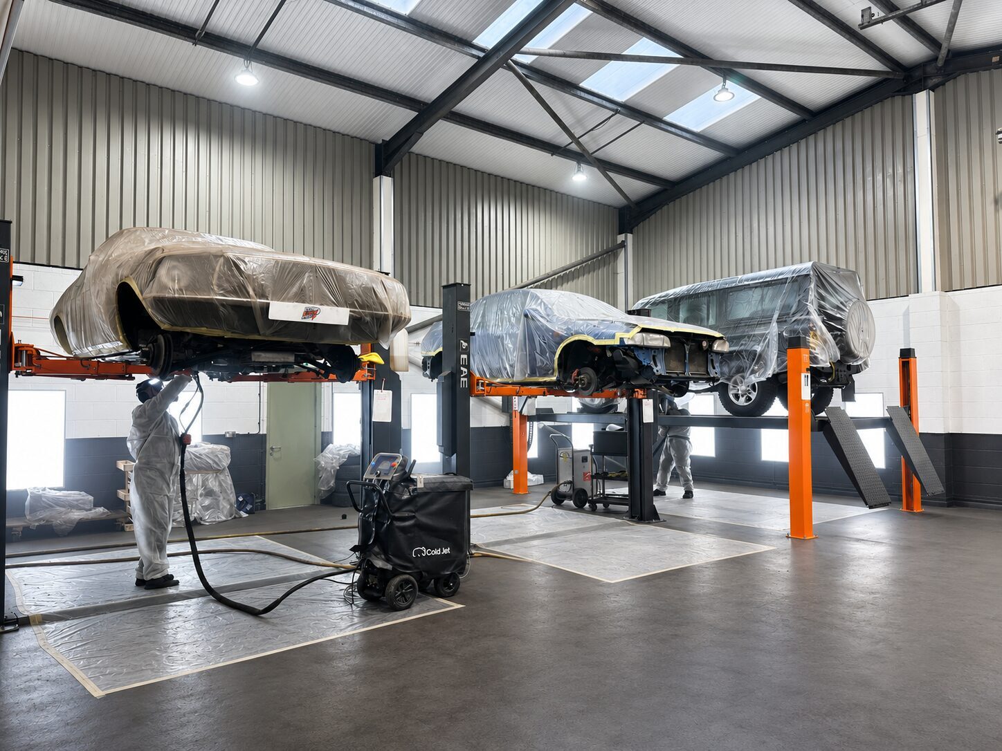 Three cars protectively wrapped and raised on workshop ramps mid-preservation, undertrays off and undersides exposed for dry ice blasting at IceBlastPro in Oxfordshire
