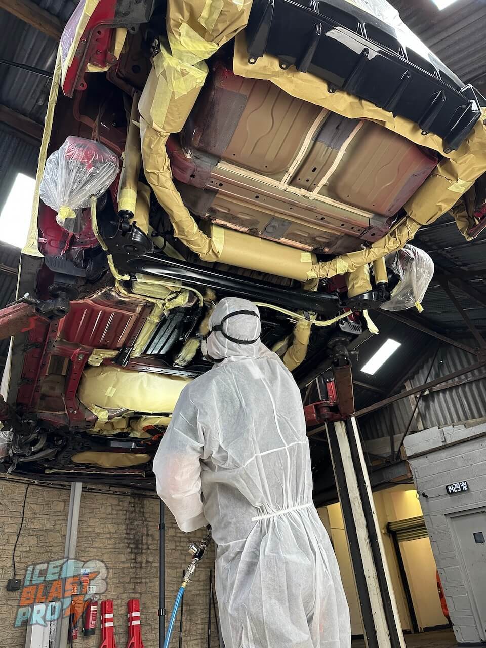 Technician in a protective suit spraying Dinitrol protection across a vehicle underbody on a ramp