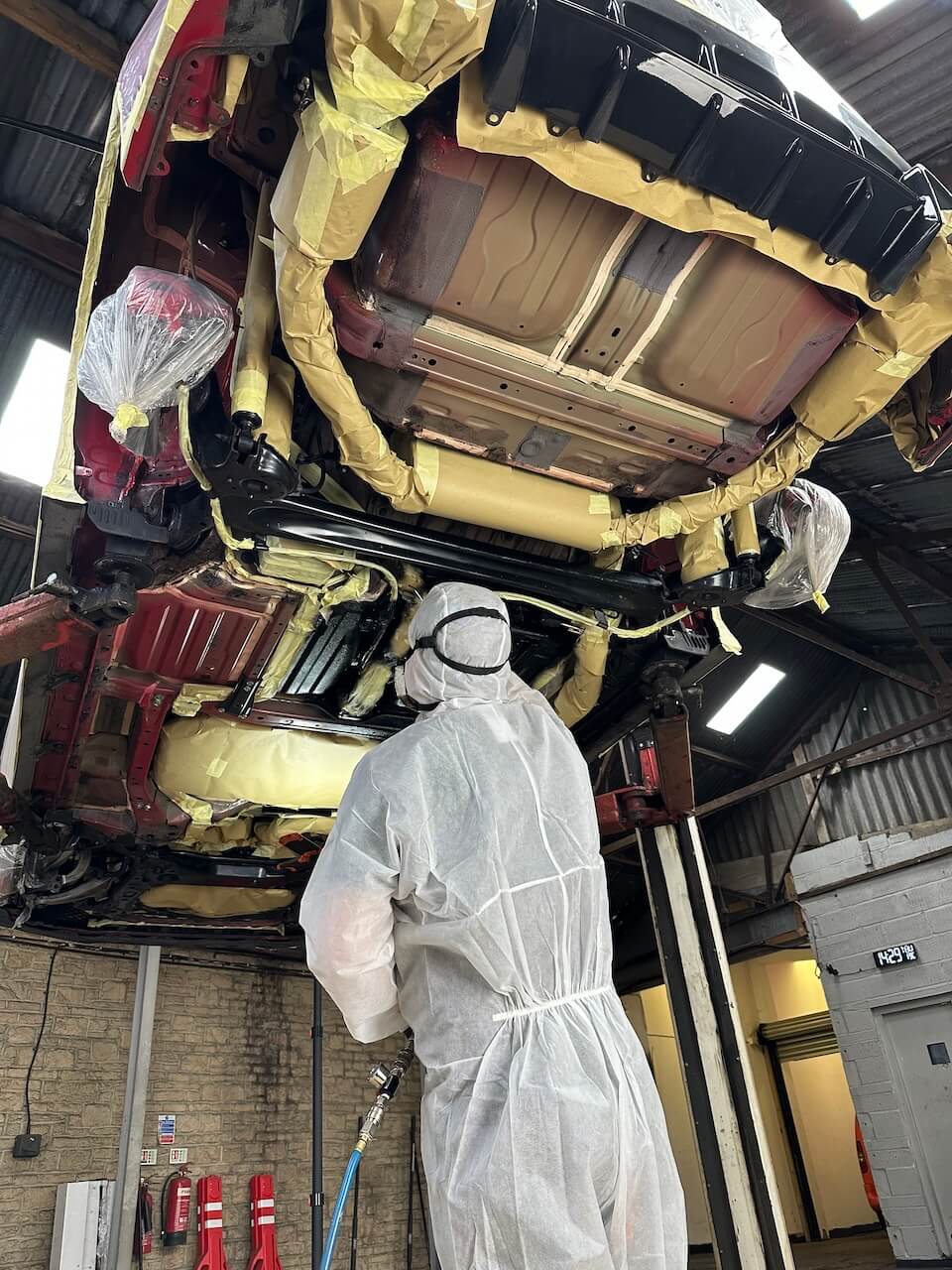 Technician in a protective suit spraying Dinitrol protection across a vehicle underbody on a ramp