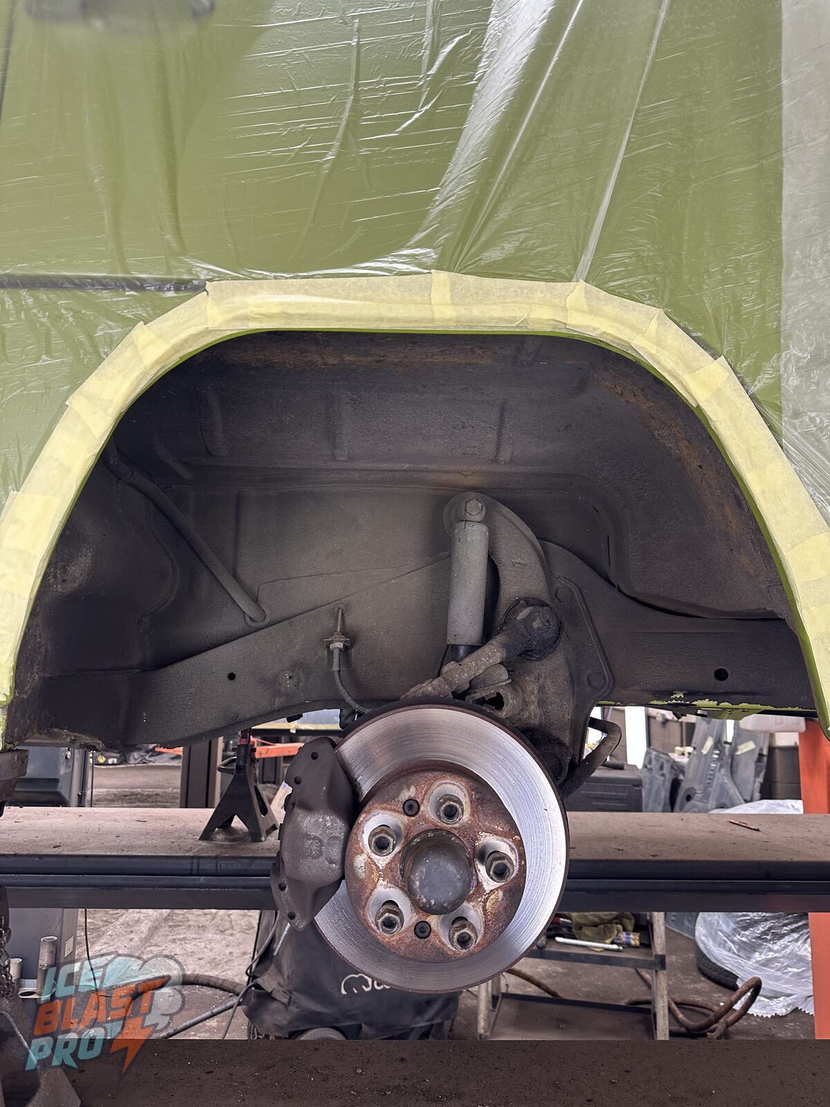 Rear wheel arch of a Volkswagen T2 camper before underseal removal, with old, grimy coating over the arch and suspension and a corroded hub and brake assembly