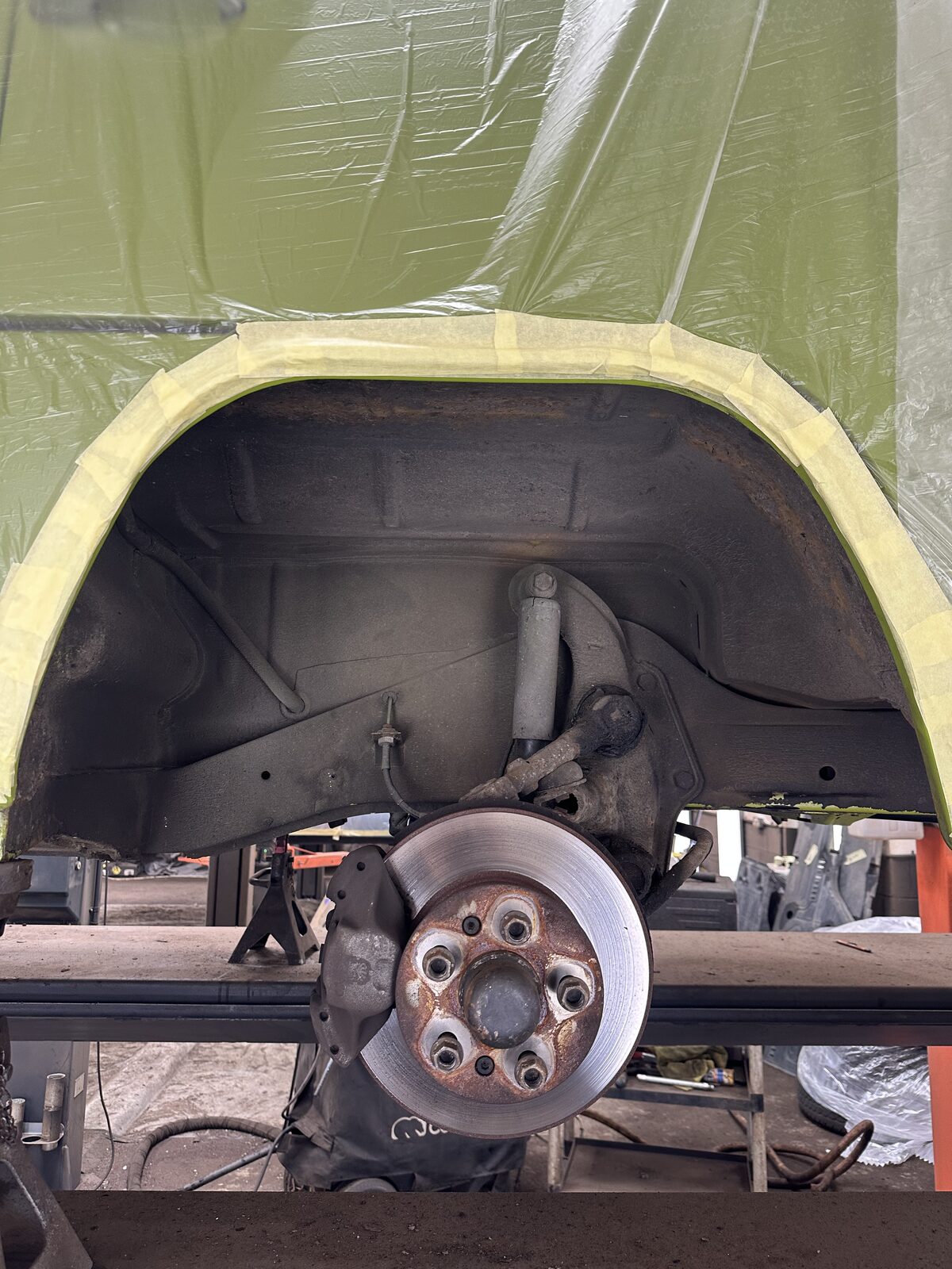 Rear wheel arch of a Volkswagen T2 camper before underseal removal, with old, grimy coating over the arch and suspension and a corroded hub and brake assembly