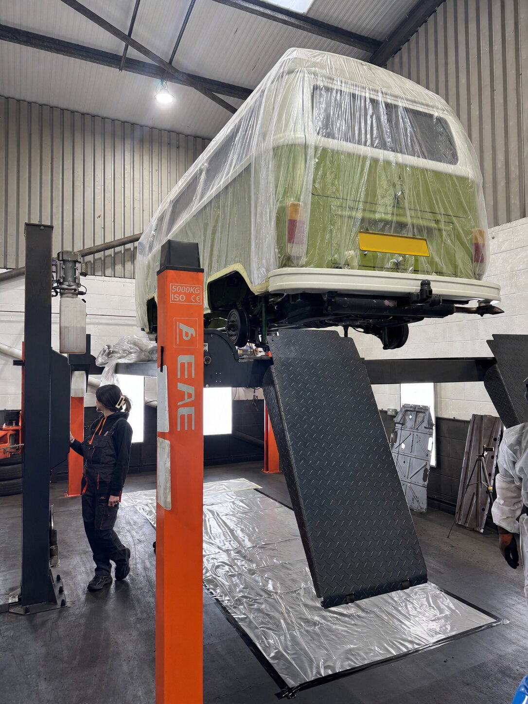 The green Volkswagen T2 camper raised on a two-post ramp with its bodywork wrapped in protective sheeting at the IceBlastPro workshop, masked and protected before any underbody work began